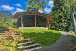 Eine hölzerne Gradieranlage steht auf einer grünen Anhöhe, umgeben von Bäumen. Ein asphaltierter Weg und eine Steintreppe führen zu ihr hinauf.