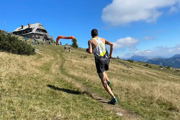 Gedenklauf zum Aflenzer Schönleitenhaus Ein Läufer läuft auf einem schmalen Bergweg über eine Almwiese bergauf. Vor ihm befinden sich eine Berghütte und weitere Teilnehmer, im Hintergrund ist eine Berglandschaft zu sehen.