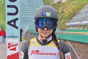 Eine Skispringerin steht mit Helm, Skibrille und Sprungski vor einer Schanzenanlage. Auf ihrer Ausrüstung sind unter anderem die Logos der Stadtwerke Trofaiach, von Stiegl und Augment zu sehen.