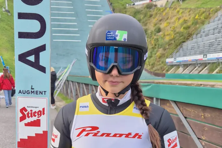 Eine Skispringerin steht mit Helm, Skibrille und Sprungski vor einer Schanzenanlage. Auf ihrer Ausrüstung sind unter anderem die Logos der Stadtwerke Trofaiach, von Stiegl und Augment zu sehen.