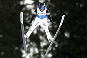 Eine österreichische Skispringerin schwebt mit weit gespreizten Skiern in der Luft. Sie trägt einen weiß-blauen Wettkampfanzug mit der Startnummer 11.