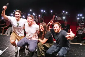 Comedy Club im Brucker Kino Drei Männer und eine Frau hocken vor Publikum auf einer Bühne