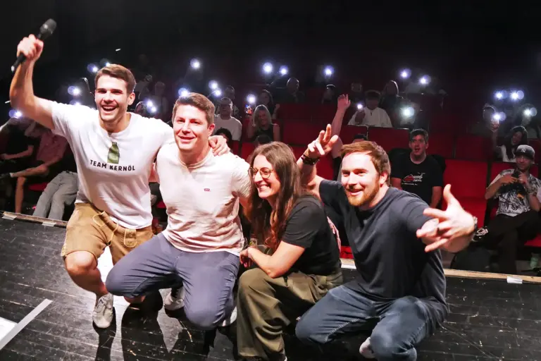 Comedy Club im Brucker Kino Drei Männer und eine Frau hocken vor Publikum auf einer Bühne