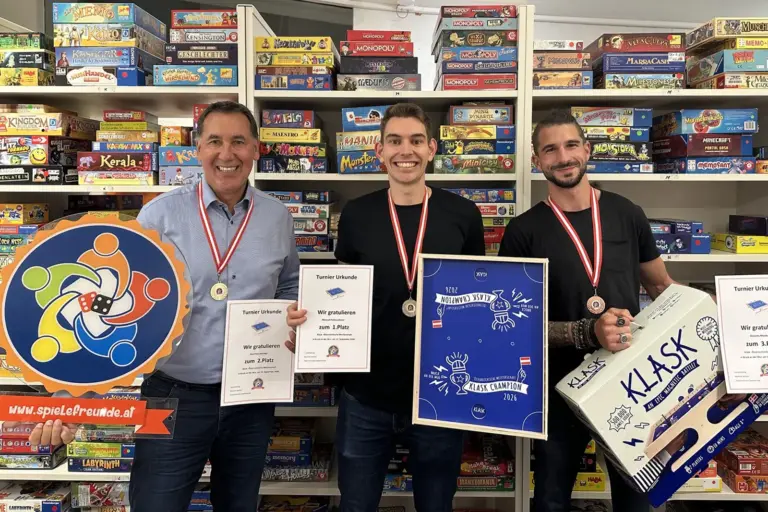 Meisterschaft der Spielefreunde in Bruck Drei Männer stehen mit Urkunden und Preisen vor einem Regal mit Spielen