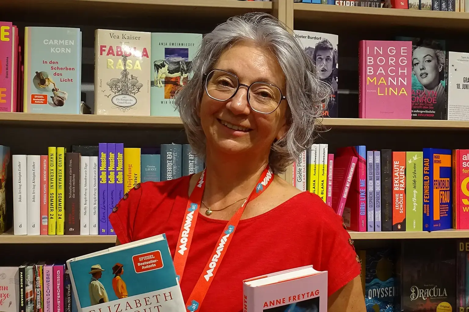 Bruck: Bücher als große Leidenschaft Eine ältere Frau steht mit zwei Büchern in der Hand vor einem Bücherregal