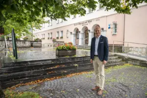 Ein Mann steht vor dem Marktgemeindeamt Thörl. Das rosafarbene Gebäude mit Rundbögen im Eingangsbereich ist von Bäumen und Blumenschmuck umgeben.