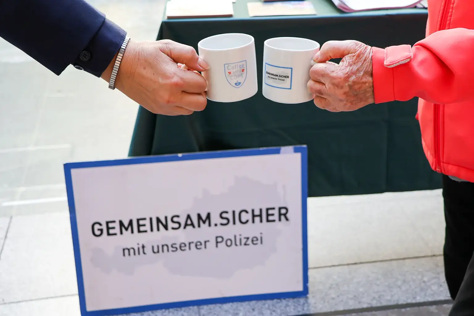 Polizei Bruck-Mürzzuschlag lädt zum Kaffeetratsch Zwei Personen stoßen mit weißen Kaffeetassen an, auf denen die Aufschriften „Coffee with Cops“ und „Gemeinsam sicher mit unserer Polizei“ zu sehen sind.