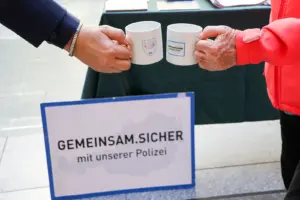 Zwei Personen stoßen mit weißen Kaffeetassen an, auf denen die Aufschriften „Coffee with Cops“ und „Gemeinsam sicher mit unserer Polizei“ zu sehen sind.