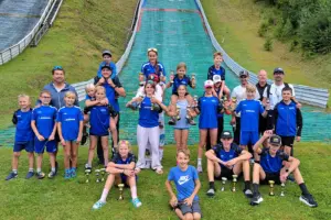 Eine Gruppe von Kindern, Jugendlichen und Erwachsenen posiert mit zahlreichen Pokalen vor einer Skisprungschanze. Viele tragen einheitliche blaue Sportbekleidung.