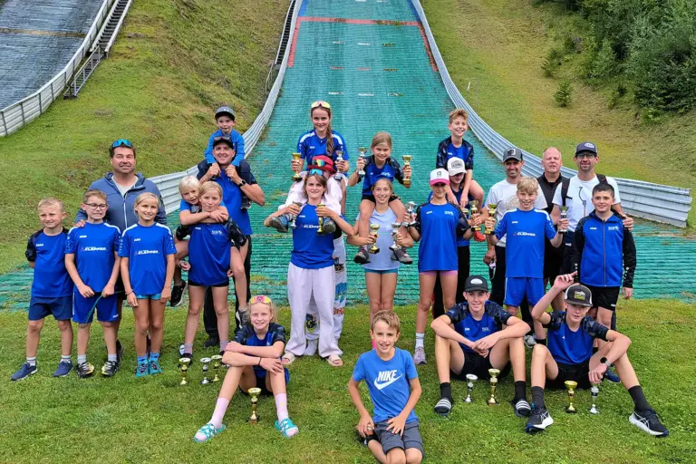 Schisprung-Landescup in Mürzzuschlag Eine Gruppe von Kindern, Jugendlichen und Erwachsenen posiert mit zahlreichen Pokalen vor einer Skisprungschanze. Viele tragen einheitliche blaue Sportbekleidung.