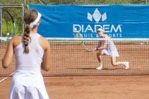 Zwei Tennisspielerinnen in weißer Sportkleidung spielen auf einem Sandplatz. Eine Spielerin steht am Netz und schlägt den Ball zurück.
