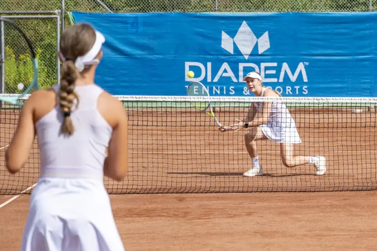 Mürztal Series: Rekordturnier beim TC Redfeld in Kapfenberg Zwei Tennisspielerinnen in weißer Sportkleidung spielen auf einem Sandplatz. Eine Spielerin steht am Netz und schlägt den Ball zurück.