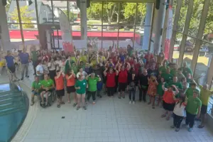 Eine große Gruppe von Menschen in bunter Sportkleidung versammelt sich in einer Schwimmhalle neben einem Becken. Viele Teilnehmer winken lächelnd in die Kamera, einige sitzen in Rollstühlen.