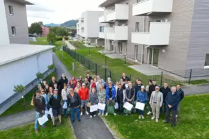 Eine große Gruppe von Menschen posiert vor einer modernen Wohnanlage mit weißen Balkonen und begrünten Außenflächen in Kindberg. Einige Personen halten Geschenktaschen in den Händen.