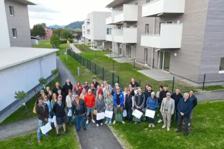 27 Wohnungen in der Kindberger Aulandgasse übergeben Eine große Gruppe von Menschen posiert vor einer modernen Wohnanlage mit weißen Balkonen und begrünten Außenflächen in Kindberg. Einige Personen halten Geschenktaschen in den Händen.
