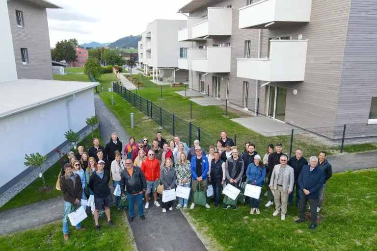27 Wohnungen in der Kindberger Aulandgasse übergeben Eine große Gruppe von Menschen posiert vor einer modernen Wohnanlage mit weißen Balkonen und begrünten Außenflächen in Kindberg. Einige Personen halten Geschenktaschen in den Händen.