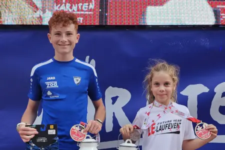 Leobener sind in Kärnten zu Siegen gelaufen Ein Jugendlicher und ein Mädchen präsentieren nach einer Laufveranstaltung lächelnd ihre Medaillen. Beide tragen Sportkleidung und stehen vor einer Veranstaltungswand.