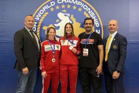 Zwei WM-Medaillen für Leobener Muaythai-Kämpferinnen Zwei Sportlerinnen in roten Trainingsanzügen präsentieren ihre Medaillen bei einer Weltmeisterschaft. Neben ihnen stehen drei Männer, im Hintergrund ist das Logo der Veranstaltung zu sehen.