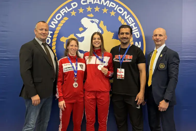 Zwei WM-Medaillen für Leobener Muaythai-Kämpferinnen Zwei Sportlerinnen in roten Trainingsanzügen präsentieren ihre Medaillen bei einer Weltmeisterschaft. Neben ihnen stehen drei Männer, im Hintergrund ist das Logo der Veranstaltung zu sehen.