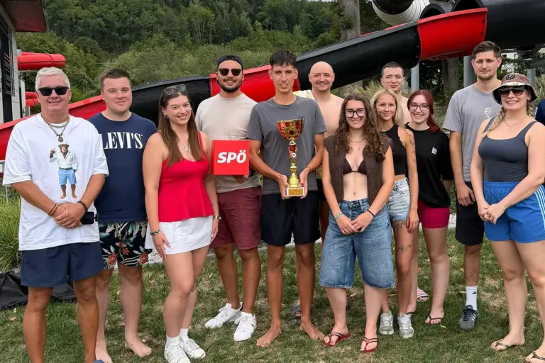 Eine Gruppe von Personen in leichter Beklediung steht mit einem Pokal auf einer Wiese