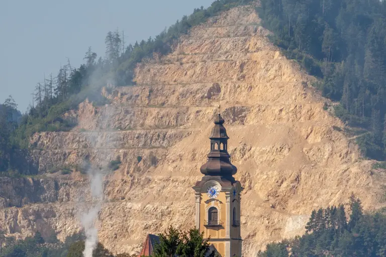 Ein Kirchturm vor einem Steinbruch auf einem bewaldeten Berg