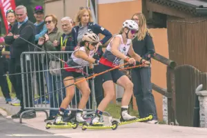 Zwei Sportlerinnen mit Helmen und Skistöcken starten auf Skirollern zu einem Rennen. Hinter ihnen verfolgen mehrere Zuschauer das Geschehen.