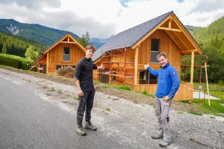 Touristisches Vater-Sohn-Projekt am Fuße des Hochschwabs Zwei Männer stehen vor zwei neu errichteten Holzhäusern in einer Berglandschaft und präsentieren die Gebäude mit ausgestreckten Armen.