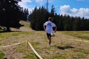 Ein Läufer in weißem T-Shirt und schwarzer Hose läuft auf einer mit Absperrbändern markierten Strecke über eine sonnige Bergwiese. Im Hintergrund erstreckt sich ein dichter Nadelwald.