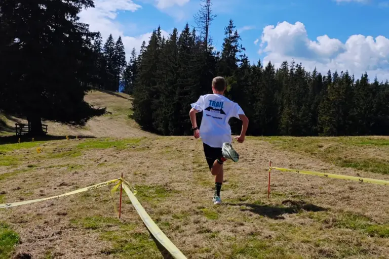 Gasener siegte beim Stanzer Trailrun Ein Läufer in weißem T-Shirt und schwarzer Hose läuft auf einer mit Absperrbändern markierten Strecke über eine sonnige Bergwiese. Im Hintergrund erstreckt sich ein dichter Nadelwald.