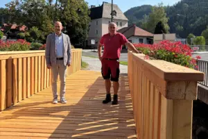 Zwei Männer stehen auf einer neuen Holzbrücke mit Blumenschmuck. Im Hintergrund sind Häuser, eine Straße und bewaldete Hügel zu sehen.