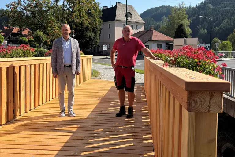 Fußgängerbrücke im Thörler Zentrum wurde neu errichtet Zwei Männer stehen auf einer neuen Holzbrücke mit Blumenschmuck. Im Hintergrund sind Häuser, eine Straße und bewaldete Hügel zu sehen.