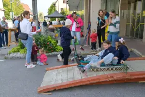 Eine Gruppe Menschen auf einem innerörtlichen Platz, im Vordergrund zwei Mädchen auf einer Rollenrodel
