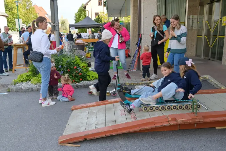 Eine Gruppe Menschen auf einem innerörtlichen Platz, im Vordergrund zwei Mädchen auf einer Rollenrodel
