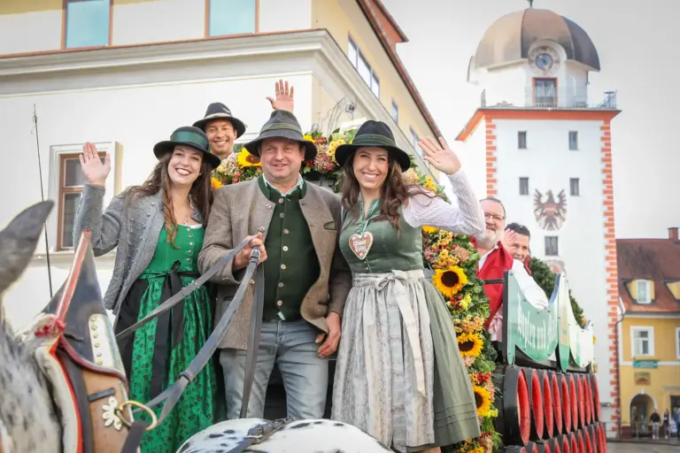 Gösser Kirtag 2026 Lächelnde und winkende Menschen in Trachtenkleidung auf einer mit Bierfässern beladenen Pferdekutsche