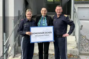 Ein Mann und eine Frau in Uniform präsentieren mit einer Frau in Zivil eine Tafel vor einer Treppe zu einem Haus
