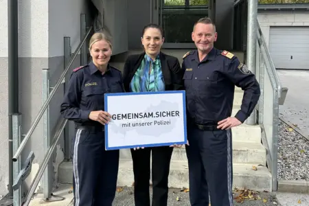 Ein Mann und eine Frau in Uniform präsentieren mit einer Frau in Zivil eine Tafel vor einer Treppe zu einem Haus