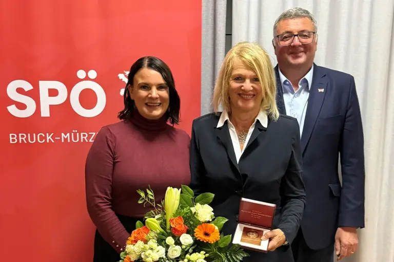 Bruck: Auszeichnung für Andrea Winkelmeier Zwei Frauen und ein Mann stehen mit einem Blumenstrauß vor einem Rollup