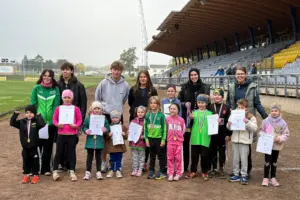 Eine Gruppe von Kindern mit Medaillen und Urkunden posiert gemeinsam mit mehreren Jugendlichen und Erwachsenen auf der Laufbahn eines Sportstadions für ein Gruppenfoto. Im Hintergrund sind das Spielfeld und die überdachte Zuschauertribüne zu sehen.