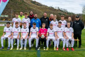 Eine Jugendfußballmannschaft in weißen Trikots und Hosen posiert gemeinsam mit mehreren Betreuern auf einem Fußballplatz für ein Gruppenfoto. Die vordere Reihe sitzt auf Bänken, die hintere steht. In der Mitte sitzt ein Tormann im pinken Trikot mit einem Fußball in den Händen. Im Hintergrund sind ein Zaun und bewaldete Hügel zu sehen.