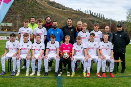 Neue Dressen für Brucker U16-Kicker Eine Jugendfußballmannschaft in weißen Trikots und Hosen posiert gemeinsam mit mehreren Betreuern auf einem Fußballplatz für ein Gruppenfoto. Die vordere Reihe sitzt auf Bänken, die hintere steht. In der Mitte sitzt ein Tormann im pinken Trikot mit einem Fußball in den Händen. Im Hintergrund sind ein Zaun und bewaldete Hügel zu sehen.