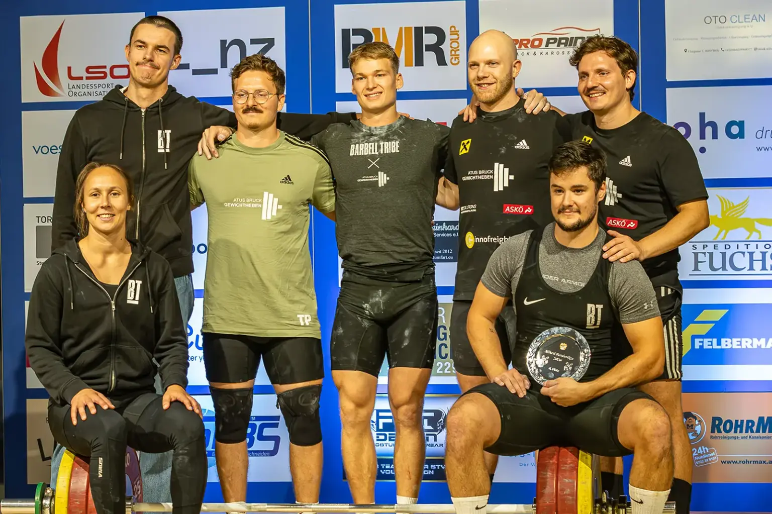 Hauchdünne Niederlage für Brucker Gewichtheber Sieben Mitglieder des ATUS Bruck Gewichtheben posieren vor einer Sponsorenwand für ein gemeinsames Gruppenfoto.