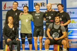 Sieben Mitglieder des ATUS Bruck Gewichtheben posieren vor einer Sponsorenwand für ein gemeinsames Gruppenfoto.