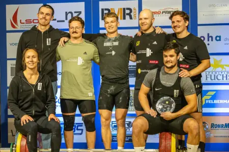 Hauchdünne Niederlage für Brucker Gewichtheber Sieben Mitglieder des ATUS Bruck Gewichtheben posieren vor einer Sponsorenwand für ein gemeinsames Gruppenfoto.