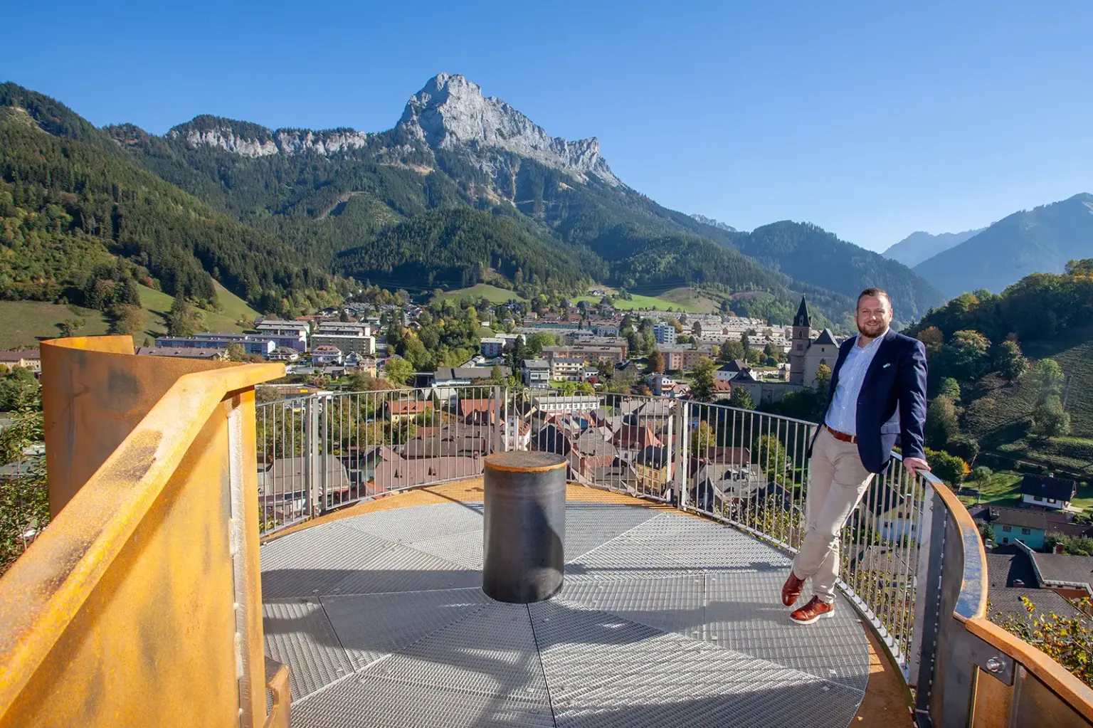 Ein Mann steht lächelnd auf einer Aussichtsplattform, im Hintergrund eine Stadt und eine Berglandschaft