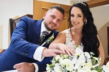 Ein Mann mit blauem Anzug und eine Frau mit weißem Kleid halten ihre rechten Hände mit jeweils einem Ring am Ringfinger über einem weißen Blumenstrauß
