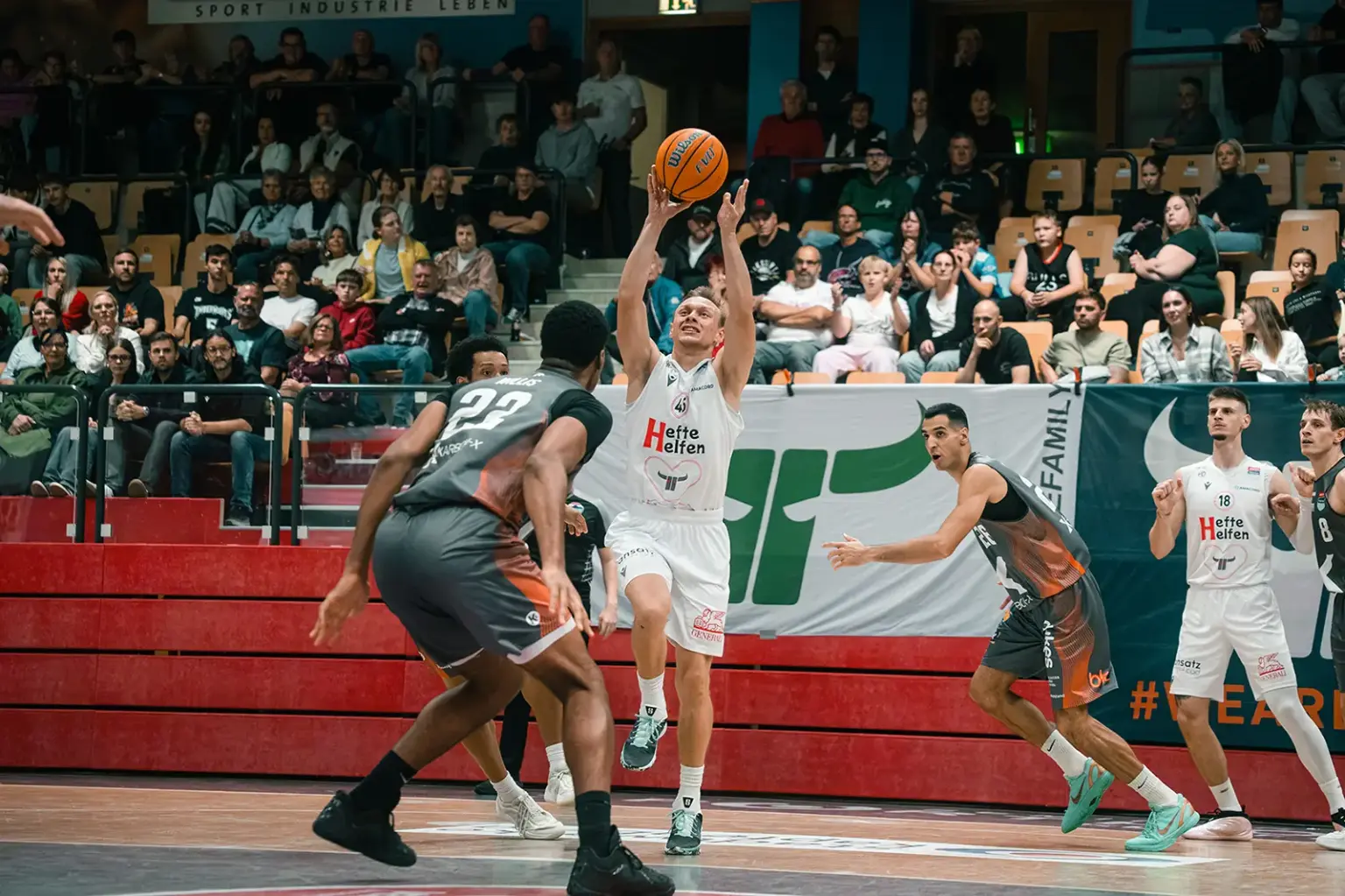 Fünfter Sieg im fünften Spiel für die Kapfenberg Bulls Ein Basketballspieler der Kapfenberg Bulls setzt vor mehreren Gegenspielern zum Korbwurf an. Im Hintergrund verfolgen zahlreiche Zuschauer die Partie.