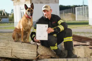 Ein Mann mit Hund sitzt auf Steinbrocken und präsentiert eine Urkunde