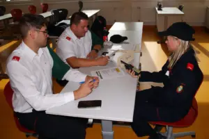 Eine junge Frau mit einem Funkgerät sitzt zwei Männern in Uniform gegenüber