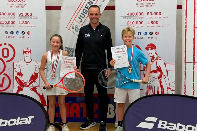 Zwei junge Tennisspieler mit Medaillen, Urkunden und Tennisschlägern stehen gemeinsam mit einem Betreuer vor den Bannern des Österreichischen Tennisverbandes.