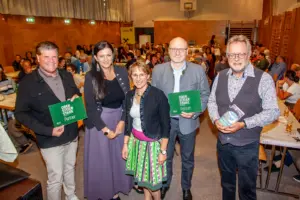Zwei Frauen und drei Männer mit Schildern und einem Buch in Händen stehen in einem Turnsaal, im Hintergrund voll besetzte Tische